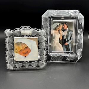 May include: Two clear glass picture frames. The square frame features a border of molded hearts. The rectangular frame features a border of molded floral and leaf patterns. The square frame holds a photo of a child, and the rectangular frame holds a photo of a bride and groom.