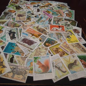 May include: A large collection of rectangular postage stamps featuring various animal illustrations, including a squirrel, rhinoceros, and dinosaur. The stamps display diverse colors, international text, and numerical values, scattered across a dark surface.