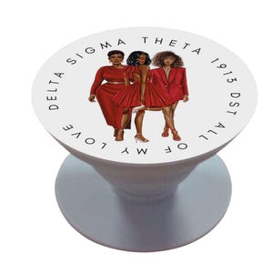 May include: A white pop-up phone grip featuring an illustration of three women in red dresses. The text "DELTA SIGMA THETA 1913 DST ALL OF MY LOVE" is printed around the edge of the grip. The grip has a white base.