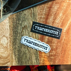 May include: Two black and white stickers with the text "Y'ALTERNATIVE" surrounded by a barbed wire design. The stickers are on a wooden surface.