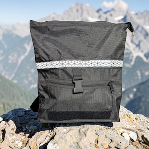 May include: Black backpack with a reflective white geometric pattern band across the top. The backpack has a front zippered pocket, a black buckle closure, and a black velcro patch. The background features a mountain range.