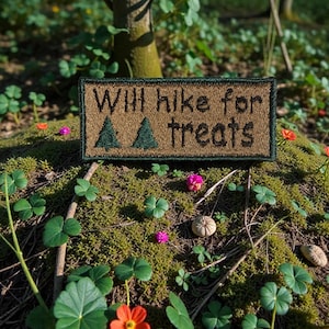May include: A rectangular patch with the text "Will hike for treats" and two green tree illustrations. The patch is brown with a green border and is set in a natural outdoor environment with green plants and flowers.