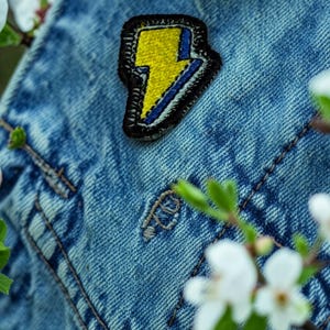 May include: A yellow and blue lightning bolt patch with a black border is sewn onto a blue denim jacket. The patch is a vibrant accent against the textured fabric. White flowers and green leaves are in the background.