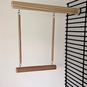 May include: A wooden bird swing with a horizontal perch and two vertical dowels suspended from a horizontal wooden bar. The swing is attached to a black wire cage. The swing is made of light brown wood and silver metal hardware.
