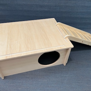 Puede incluir: Una casa de madera de color claro para mascotas con un techo inclinado y una escalera adjunta. La casa tiene una entrada circular y está diseñada para animales pequeños. La madera tiene un acabado natural.