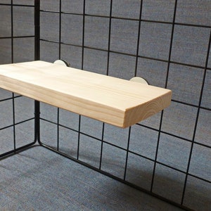 May include: A light-colored wooden shelf, rectangular in shape, is mounted on a black wire grid. The shelf is supported by two small, round, silver-colored brackets. The grid is set against a gray background.