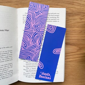Op de afbeelding: Twee rechthoekige bladwijzers met abstracte roze en blauwe ontwerpen. Eén heeft een patroon van golvende lijnen, de andere een blauwe achtergrond met roze vormen en de tekst "Juuls Designs."