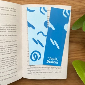 Op de afbeelding: Twee blauwe bladwijzers met abstracte vormen en witte contouren. De bladwijzer aan de linkerkant heeft een lichtblauwe achtergrond en de bladwijzer aan de rechterkant heeft een donkerblauwe achtergrond. De bladwijzer aan de rechterkant heeft de tekst "*Juuls* Designs" erop gedrukt.