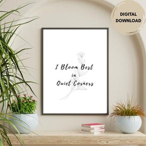 May include: A framed print with the text "I Bloom Best in Quiet Corners" in black script. The print features a delicate floral illustration. A "DIGITAL DOWNLOAD" sticker is in the upper right corner. The print is displayed on a wooden shelf with plants.