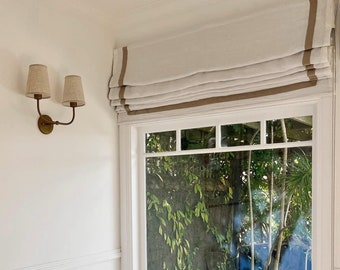 May include: A white linen roman shade with a brown trim hangs in a window with a view of lush green foliage. A white wicker side table sits next to a beige sofa with blue and white throw pillows.
