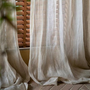 May include: Two white sheer curtains hang in front of a window with wooden blinds. The curtains are gathered at the bottom and the fabric is slightly wrinkled.