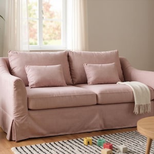 May include: A two-seater sofa in a soft pink hue, featuring two large back cushions and two smaller throw pillows. A cream-colored throw blanket with fringe rests on the sofa. The sofa sits on a black and white striped rug.