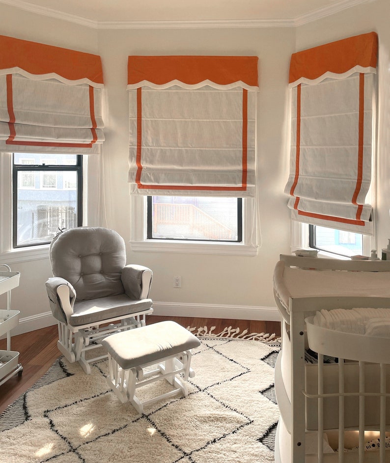 May include: A white glider rocking chair with a gray cushion and a matching footstool. The chair is in a nursery with white walls and orange and white roman shades on the windows. There is a white changing table in the background.