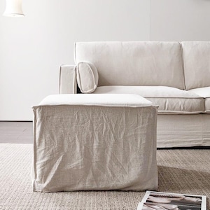 Könnte beinhalten: Ein quadratischer Hocker, bezogen mit einem hellbeigen, strukturierten Stoff im Hussen-Stil. Der Hocker weist sichtbare Nähte an den Kanten und eine leicht knittrige Oberflächenstruktur auf und steht vor einem passenden hellbeigen Sofa.