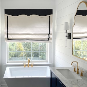 Può includere: Un bagno bianco con una vasca da bagno bianca, un lavandino bianco e uno specchio dorato con cornice nera. Il bagno ha pareti in legno bianco e una finestra bianca con una tenda romana bianca e nera.