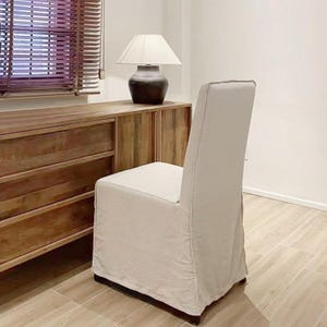 Puede incluir: Silla de comedor de color crema con respaldo alto y funda. La silla está frente a una cómoda de madera y una lámpara con pantalla blanca. La habitación tiene persianas de madera y suelo de baldosas.