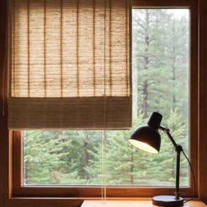 May include: A window with a natural woven shade, partially lowered, revealing a view of a lush green forest. A black desk lamp with an adjustable arm sits on a wooden surface, illuminated.