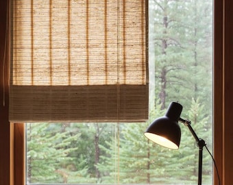May include: A window with a natural woven shade, partially lowered, revealing a view of a lush green forest. A black desk lamp with an adjustable arm sits on a wooden surface, illuminated.