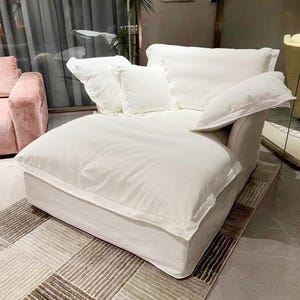 May include: A large, white, overstuffed lounge chair with a slipcovered design, featuring a wide seat, a low backrest, and three matching white throw pillows. The chair has a soft, plush appearance with visible seams along the edges of the fabric.