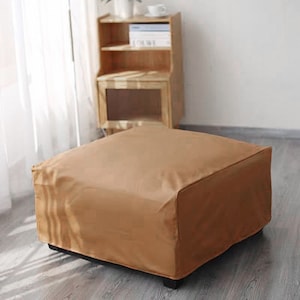 May include: A square ottoman featuring a solid tan fabric cover with clean, straight edges and a low-profile design. The piece is constructed with a structured form and sits on small, dark, block-style feet, presenting a uniform and smooth surface.