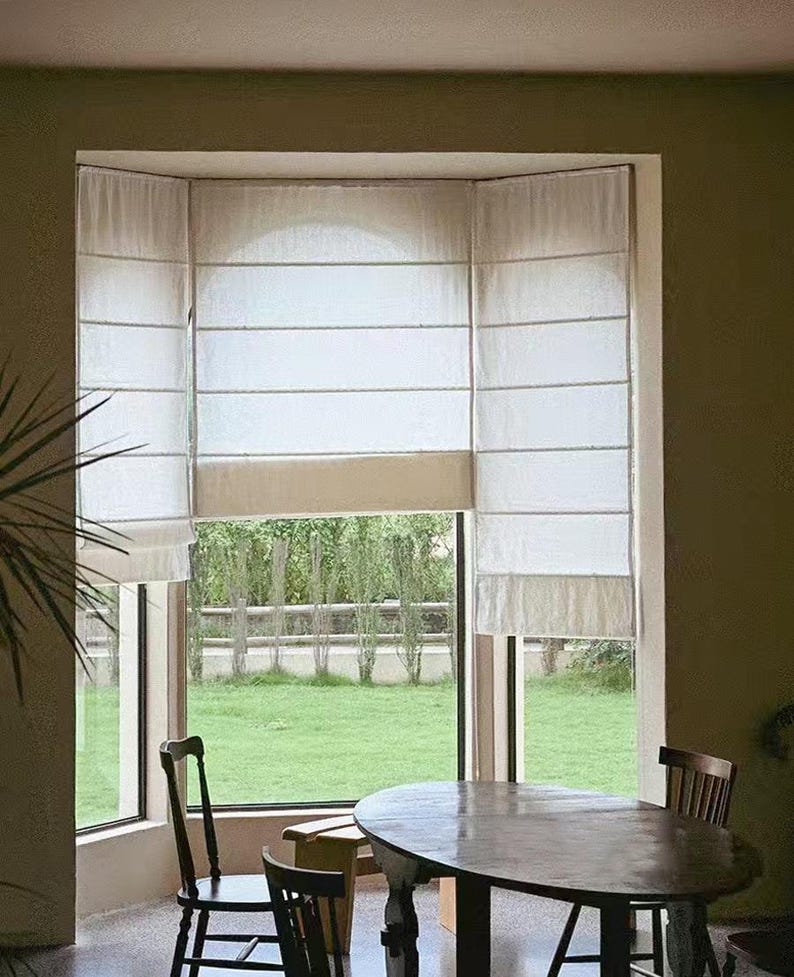 May include: A bay window with white roman shades and a view of a green lawn. A wooden table with four chairs is in the foreground.