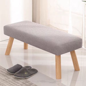 May include: A rectangular bench featuring a light gray fabric upholstered seat and four light-colored wooden legs. The bench has a clean, simple design with a flat top surface. A pair of gray fabric slippers are positioned near the bench legs.