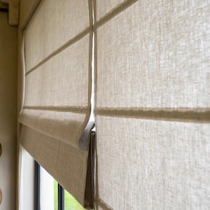 May include: Close-up of a white linen roman shade with horizontal pleats. The shade is partially raised, revealing a window with green foliage outside.