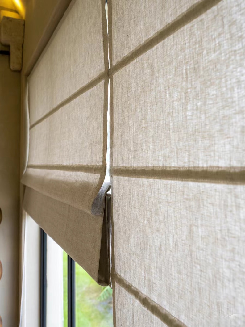 May include: Close-up of a white linen roman shade with horizontal pleats. The shade is partially raised, revealing a window with green foliage outside.