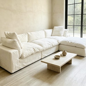 May include: A large, off-white modular sectional sofa with plush, deep-seated cushions and a matching ottoman. In front of the sofa sits a low, rectangular beige stone coffee table holding a small ceramic bowl and a rounded ceramic vase.