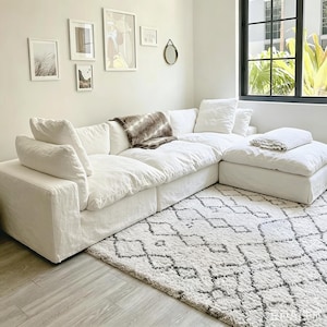 May include: A large, modular white sectional sofa with multiple cushions and a matching ottoman. A patterned rug in shades of white and gray sits beneath the sofa. Decorative framed art and a small mirror adorn the wall.