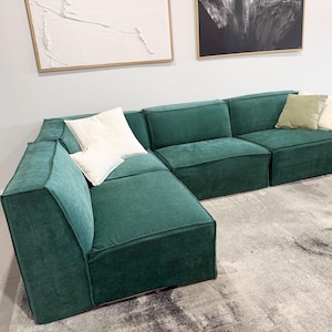 May include: A modular sectional sofa upholstered in deep emerald green fabric. The sofa features a low-profile design with wide, square armrests and back cushions. It is accented with two white square pillows and two smaller sage green pillows.