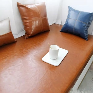 May include: A brown leather bench with two pillows, one brown and one blue, and a white cup and saucer on a white coaster.