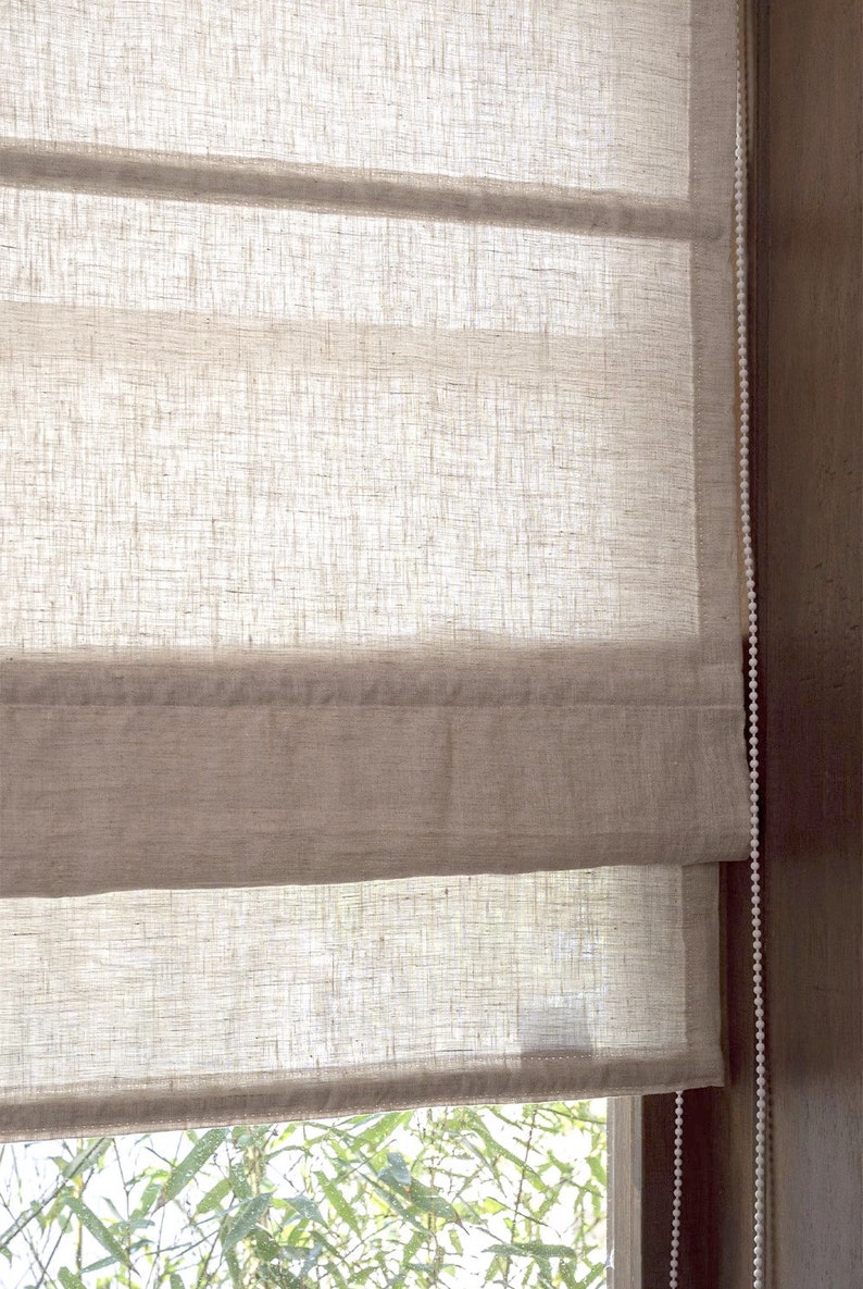 May include: A white linen Roman shade with a beige bottom border. The shade is partially raised, revealing a view of green foliage through a window.