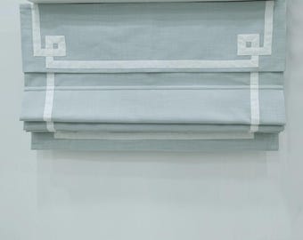 May include: A light blue roman shade with white trim and a geometric design. The shade is fully extended.