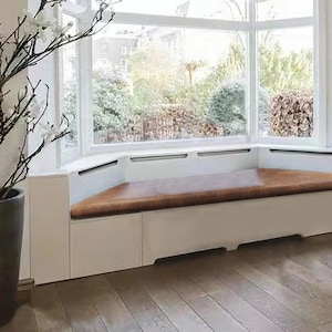 May include: A white window seat with a brown cushion. The seat is built into a bay window with white trim. The window has a view of trees and greenery.