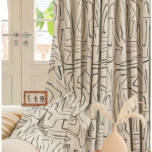 May include: White curtains with a black abstract geometric pattern. The curtains are hung in a living room with a couch, a vase with dried flowers, and a wooden table.