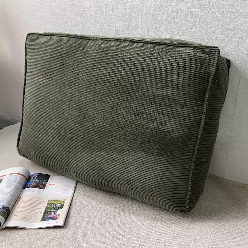 Puede incluir: Un coj&iacute;n grande de pana verde oliva con forma rectangular. El coj&iacute;n tiene una textura acanalada y un aspecto suave. Una revista est&aacute; abierta en la esquina inferior izquierda.