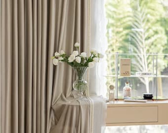 May include: A pair of beige velvet curtains with a white lace trim and a beaded edge. The curtains are partially drawn, revealing a window with a view of green trees. A vase of white flowers sits on a table in front of the window.