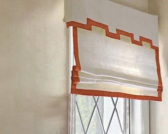 May include: White roman shade with a red trim. The shade is raised and the window behind it has a diamond pattern.