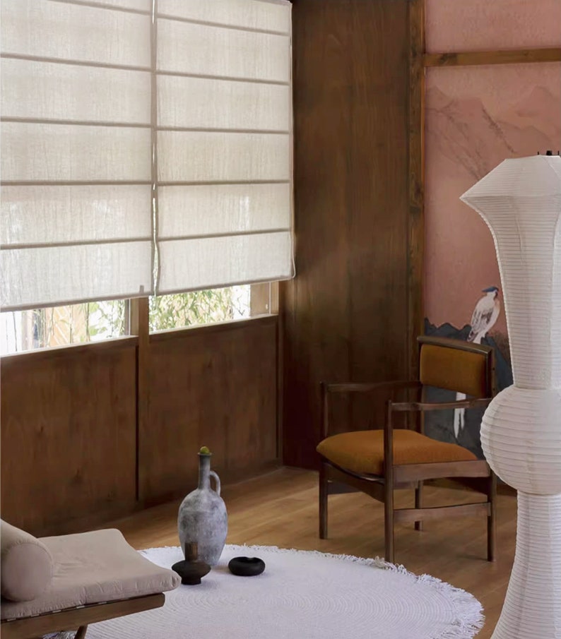 May include: A white linen Roman shade hangs in a window with a wooden frame. A brown leather chair with a wooden frame sits in the corner of the room. A white ceramic vase with a green plant sits on the floor next to a white rug.