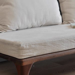 Könnte beinhalten: Ein hellbeiges Leinen-Sofa mit einem Holzrahmen. Das Sofa hat zwei Kissen und ein Buch auf der Sitzfläche.