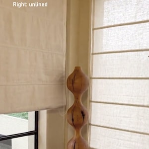 May include: Two white linen Roman shades with blackout liner on the left and unlined on the right. The shades are partially raised, revealing a window with a view of green foliage. A wooden sculpture with a bulbous shape is positioned in the corner of the window.