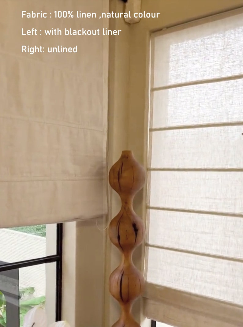 May include: Two white linen Roman shades with blackout liner on the left and unlined on the right. The shades are partially raised, revealing a window with a view of green foliage. A wooden sculpture with a bulbous shape is positioned in the corner of the window.
