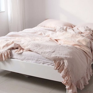 May include: A blush pink linen duvet cover with ruffled edges. The duvet is on a white bed frame and is slightly rumpled.