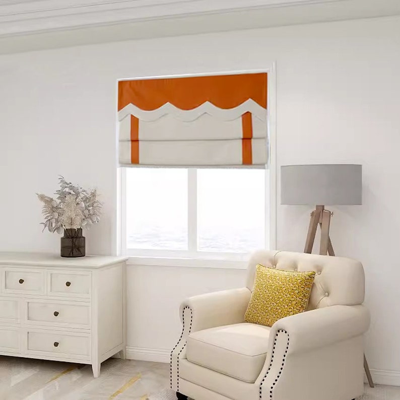May include: A white armchair with a yellow patterned pillow sits in a room with a white dresser and a window with orange and white roman shades.
