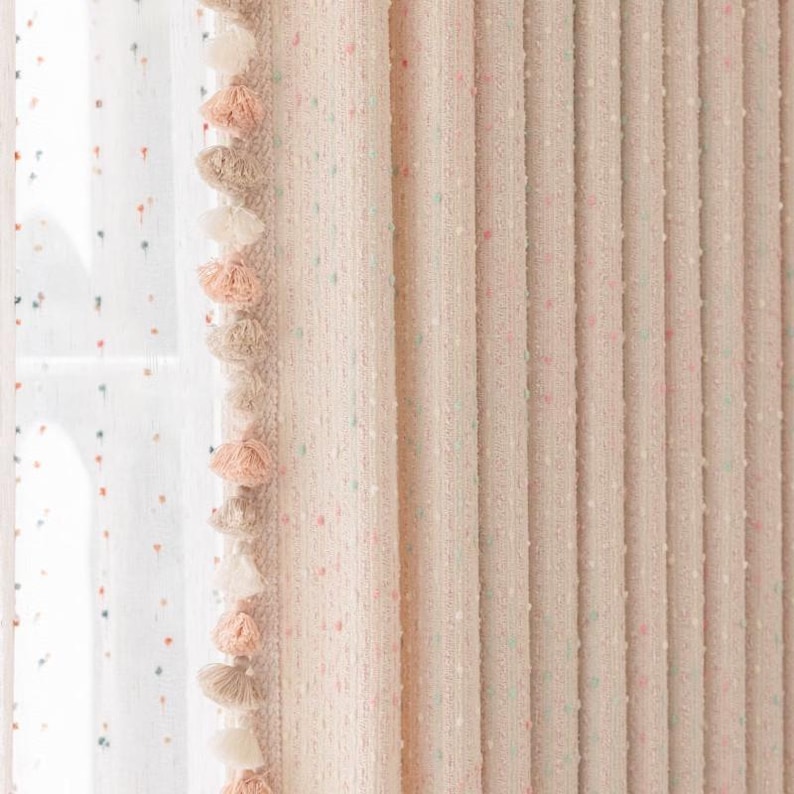 May include: A pair of pink curtains with a white background and small colorful dots. The curtains have a fringe trim with tassels in shades of pink, white, and beige.