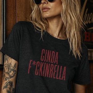 May include: A dark charcoal gray short-sleeved t-shirt featuring the red text CINDA F*CKINRELLA printed on the front. The shirt has a crew neckline and a standard fit, showcasing a bold graphic design with a distressed texture on the lettering.