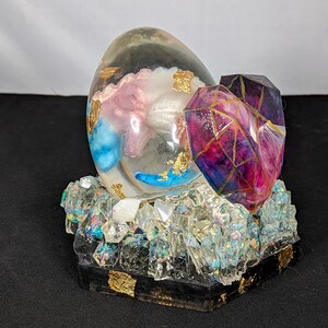 Puede incluir: Una escultura decorativa de resina con forma de huevo con detalles en azul, rosa y dorado, junto con una pieza facetada en forma de corazón. La escultura descansa sobre una base de cristales transparentes y resina negra.