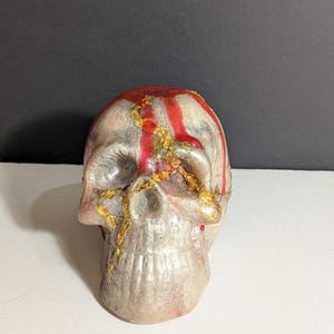 May include: A decorative skull with a gray and white base, adorned with red stripes and gold accents. The skull has a detailed, realistic design, perfect for Halloween or gothic decor.