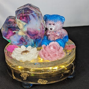 Puede incluir: Una caja decorativa de resina con un osito de peluche azul y rosa, dos estructuras en forma de cristal y detalles florales. La caja está adornada con pan de oro y luces de hadas, creando una exhibición caprichosa y artística.